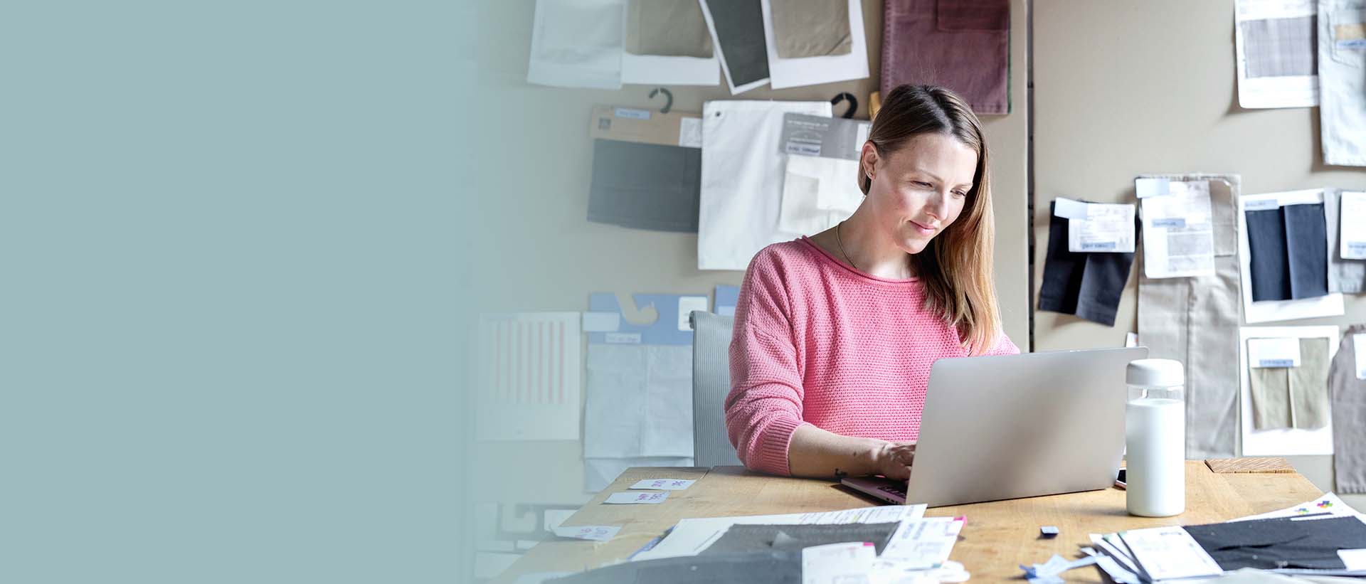 A woman is sitting at a table working on her computer. On the table and on the wall behind her there are notes and fabric samples in different colors.